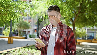 Cool hispanic man stands in sunny park, expression serious as he texts on smartphone, immersed in his relaxed but tech-driven Stock Photo