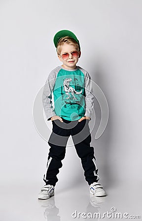 Cool kid boy in grey sweatshirt, black sport pants with stripes and white sneakers stands holding hands in pockets Stock Photo