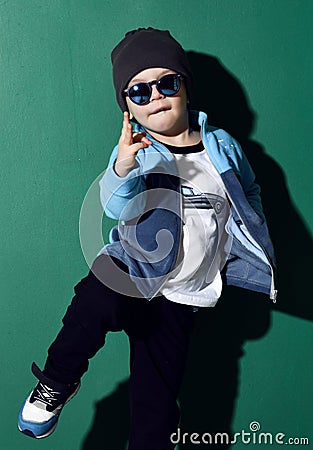 Cool kid boy in blue sunglasses, headwear, fleece jacket, pants and sneakers is dancing, showing cool sing on green Stock Photo