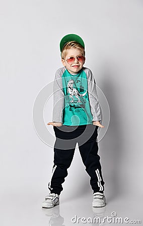 Cool kid boy in grey sweatshirt, black sport pants with stripes and white sneakers stands shrugging his shoulders Stock Photo