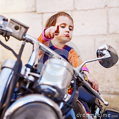 Cool little biker girl playing and having fun on fashioned motorcycle. Humorous portrait of child points to the road with finger Stock Photo