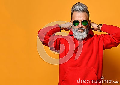 Cool old man with a gray beard is trying to put on a hood of a red sweatshirt hoodie on yellow in sunglasses Stock Photo