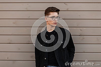 Cool serious young man hipster in fashionable denim clothes in stylish glasses is resting near wooden building on street. Handsome Stock Photo