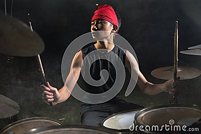 Cool and talented Asian American mixed ethnicity young boy playing drums in headband practicing and performing song in dark foggy Stock Photo