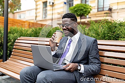 Corporate african american executive in formal wear sitting on bench near office center with laptop and takeout coffee Stock Photo