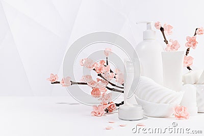 Cosmetic products in white bottles, branch of spring pink sakura flowers, toiletry for makeup facial skin care in white interior Stock Photo