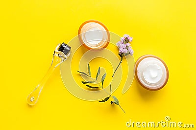 Cosmetologist tools. Dermaroller, creams near leaves and flowers on yellow background top-down flat lay Stock Photo