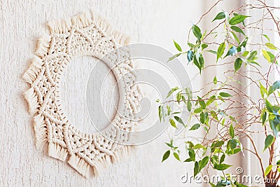 Cotton macrame mandala wall decoration hanging on white wall with green leaves. Handmade macrame wreath. Natural cotton thread. Stock Photo