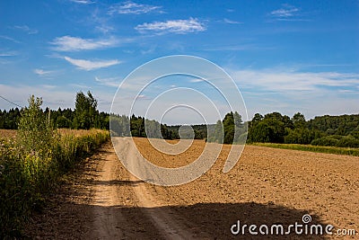 Country road along the edge of an agricultural field after plowing, rural landscape with cirrus clouds Stock Photo