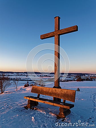 Wooden cross and bench on hill in snow-covered countryside by sunset light Stock Photo