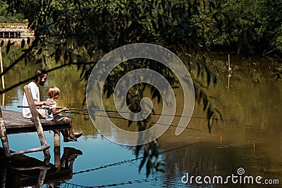 The countryside nature, trees and lake in the green colors. Father and son sitting on a wooden bridge and waiting for a Stock Photo