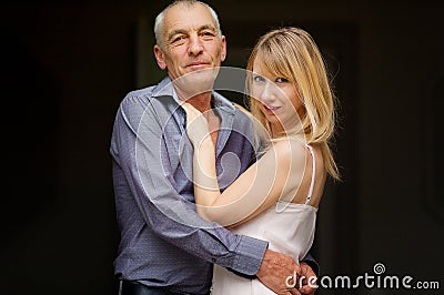 Couple with Age Difference Hugging on Black Background. Attractive Young Woman in Dress and Senior Man in Blue Shirt Stock Photo