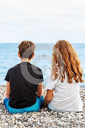 Couple of beautiful teens, first love. Guy hugs a girl sitting on the pebble beach next to each other and looking at the sea. Stock Photo