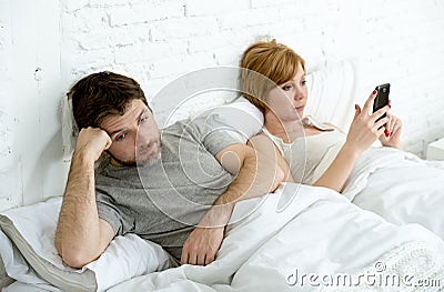 Couple in bed husband frustrated upset and unsatisfied while his internet addict wife is using mobile phone Stock Photo
