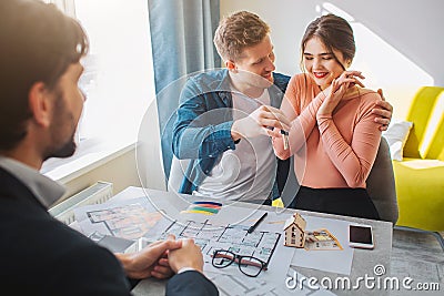 Couple buy or rent apartment together. Happy young woman get keys from man. He embrace her. Reltor sit in front of them Stock Photo