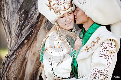 Couple in ethnic costumes embrace on background of textured wood, groom kisses bride at cheek. Stock Photo