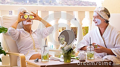 A couple of female friends share the relaxing moment taking care of their face. Sitting outdoors with a cucumber-based juice Stock Photo