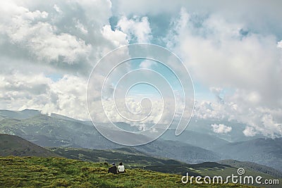 Couple hikers sitting on the mountain slope and looking at the beautiful cloudy mountain landscape Editorial Stock Photo