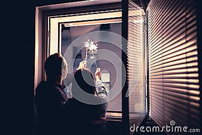 Couple holding sparklers out of the window at night. New year`s eve celebration, anniversary, party or date at home. Stock Photo