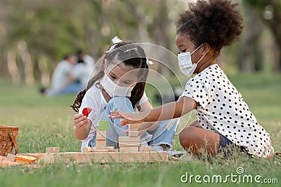 Couple little girls African and Caucasian kids wear face mask while sitting and playing wooden blocks toy in green park together Stock Photo
