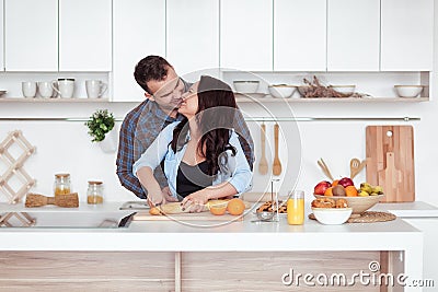 Couple making fresh organic juice in kitchen together. A young woman in a blue shirt slices a baguette. A man is hugging Stock Photo