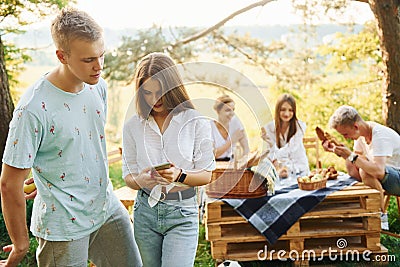 Couple with phone against people at background. Group of young people have vacation outdoors in the forest. Conception of weekend Stock Photo