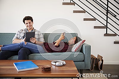 Couple Relaxing On Sofa At Home Using Mobile Phone And Digital Tablet Stock Photo