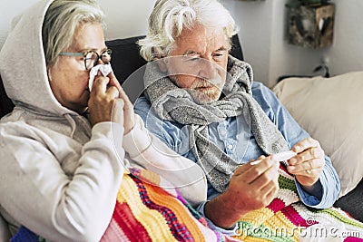 Couple of sicks seniors and mature people sitting at the sofa with fever looking at the thermometer with high temperature - two Stock Photo