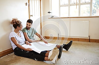Couple Sitting On Floor Looking At Plans In Empty Room Of New Home Stock Photo