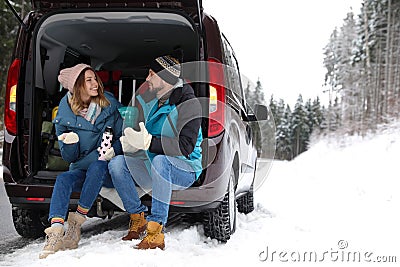 Couple sitting in open car trunk full of luggage near road, space for text. Winter Stock Photo