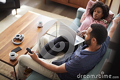 Couple Sitting On Sofa At Home Using Laptop Computer And Watching TV Stock Photo