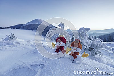 Couple two little snowmen in red hat and scarf on snowy field. Beautiful winter sunset background. Landscape with high mountains. Stock Photo