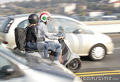 Couple wearing Italian flag helmets riding gray scooter over the city street bridge in busy traffic in motion blur Editorial Stock Photo