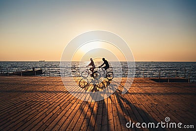 Couple of young hipsters cycling together at the beach at sunrise sky at wooden deck summer time Stock Photo