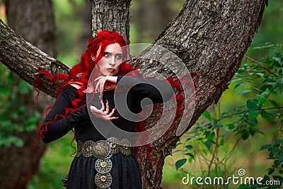 Courageous young lady with long red hair in image of fabulous historical character of witch and priestess in a mystical forest Stock Photo