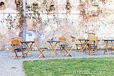 Courtyard outdoor cafe area with wooden tables and chair, green grass lawn and ruined shbby brick wall on the background. Open air Stock Photo