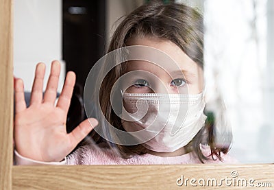 COVID-19 coronavirus concept, little girl in face mask looking through window at home or clinic. Portrait of sad kid during Stock Photo