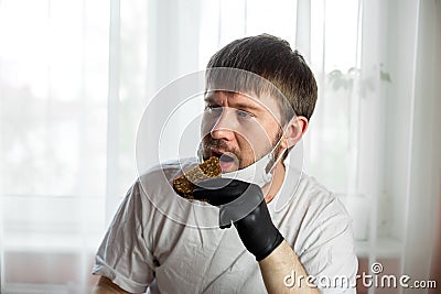 COVID-19. Young caucasian man in white mask and black gloves with a slice of bread in his hands in the kitchen during a pandemic. Stock Photo