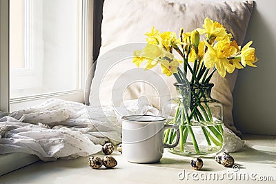 Cozy Easter, spring still life scene. Mug of coffee, wooden plate, quail eggs and vase of flowers on windowsill. Floral Stock Photo