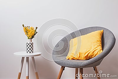 Cozy grey chair with yekllow pillow and flowers in the vase standing near the white wall. Stock Photo