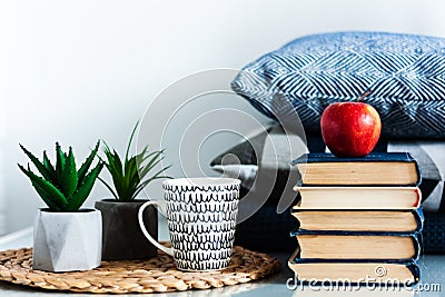 Cozy home interior decor: cup of coffee, stack of books, red apple, plants in pots on a wicker stand, blue pillows on a white Stock Photo