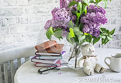 Cozy home still life - bouquet of lilacs, stack of books, glasses, toy bear, mug on the table in a bright room Stock Photo