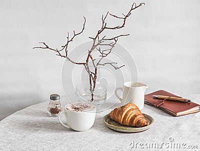 Cozy morning - cappuccino, croissant, vase with branches, leather planner notebook on the table in a bright cozy room. Cozy home Stock Photo