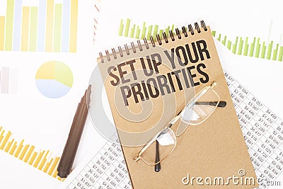 In a craft colour notebook is a Set up your priorities inscription, next to pencils, glasses, graphs and diagrams Stock Photo