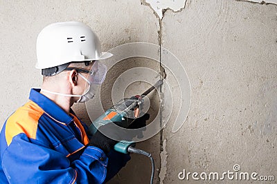 A craftsman in a blue building suit, helmet and glasses repairs a wall with a power tool Stock Photo