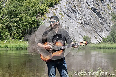 crazy man musician, guitarist on vacation. 23 year guy with beard in black shirt, cap, sunglasses holds steaming guitar Stock Photo