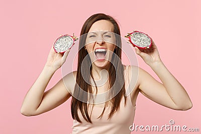 Crazy screaming young woman holding two halfs of fresh ripe pitahaya, dragon fruit isolated on pink pastel wall Stock Photo