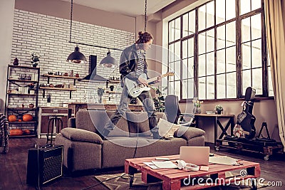 Crazy young rock musician standing on sofa and playing music Stock Photo