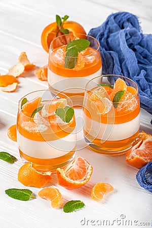 Creamy panna cotta with orange jelly in beautiful glasses, fresh ripe mandarin, blue textile on white wooden background. Delicious Stock Photo