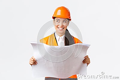 Creative asian female architect imaging her plan, standing in safety helmet and looking away thoughtful while reading Stock Photo